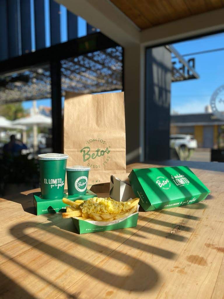 Packaging de Betos Lomitos sobre una mesa al sol: bolsa, vasos, caja y papas